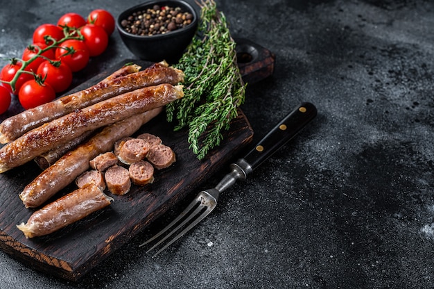 Salchicha de carne asada sobre una tabla de madera con hierbas y tomate. Fondo negro. Vista superior. Copie el espacio.