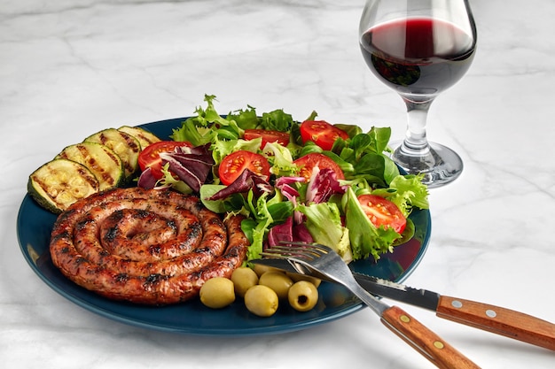 Salchicha a la parrilla con tomates picados y hierbas en una placa azul con una copa de vino tinto sobre una mesa de luz