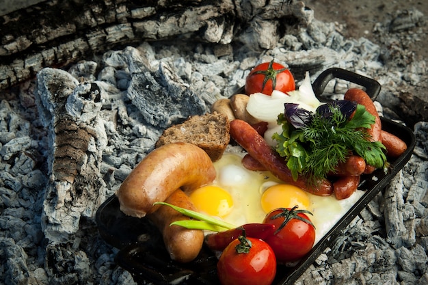 Las salchichas ahumadas con tomates y huevos se encuentran sobre carbón. el plato se cuece y se ahuma sobre carbón