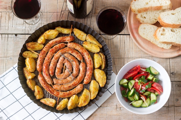 Foto salchichas de carne aromática con patatas, ensalada y vino en una mesa de madera.