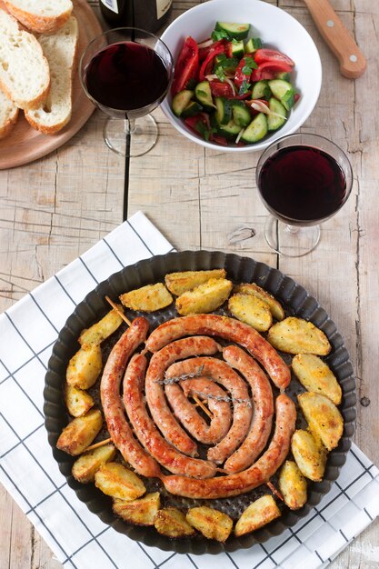 Salchichas de carne aromática con patatas, ensalada y vino en una mesa de madera.