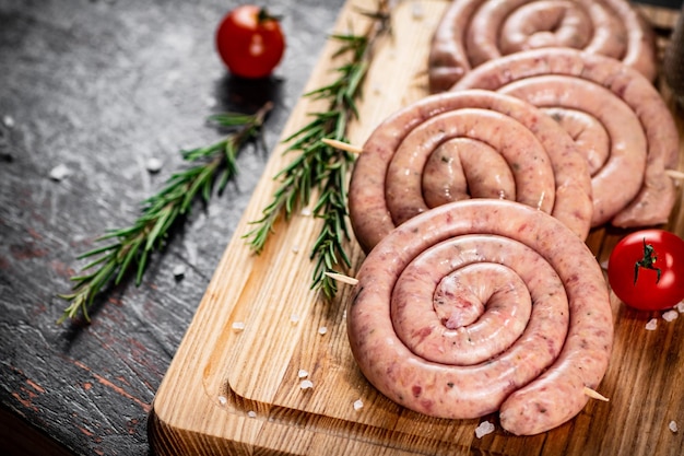 Salchichas crudas en una tabla de cortar con una ramita de romero y tomates