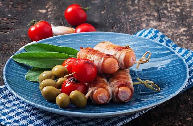 Salchichas a la parrilla envueltas en tiras de tocino con tomates y hojas de salvia