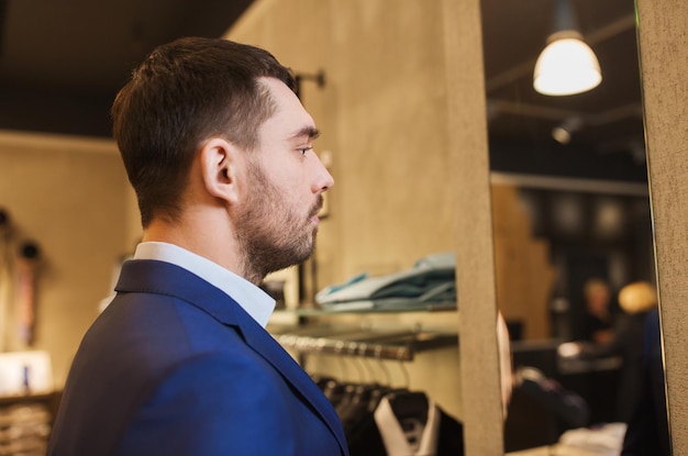 Sale, Shopping, Fashion, Style und People Concept - eleganter junger Mann in Anzugjacke beim Spiegeln im Einkaufszentrum oder Bekleidungsgeschäft