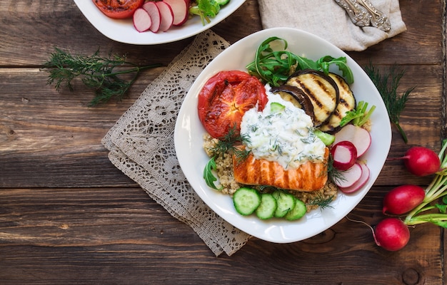 Salmão grelhado, berinjela e tomate com molho de quinua e tzatziki em madeira rústica. Jantar saudável.