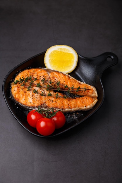 Salmón a la parrilla con tomates cherry, tomillo y limón en una pequeña sartén negra. El pescado frito es un plato tradicional de la cocina casera y los restaurantes. Enfoque selectivo de fondo negro.