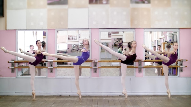 Foto en el salón de baile, jóvenes bailarinas con leotardes púrpuras realizan una actitud de eliminación en zapatos de punta.