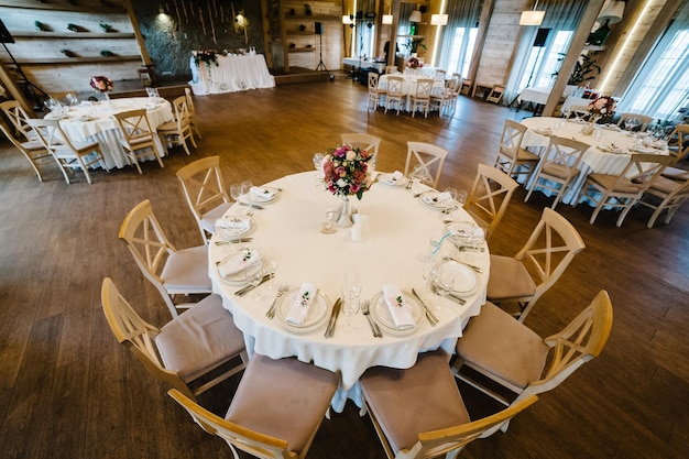 En el salón de banquetes de bodas, se colocan sillas y mesas festivas para invitados cubiertas con un mantel y decoradas con una composición de flores y vegetación Fiesta de bodas en carpa con paredes de madera de estilo