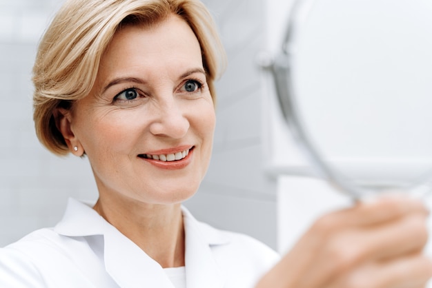 En el salón de belleza. Retrato recortado de mujer mayor en bata blanca de laboratorio mirando el espejo de mano y sonriendo mientras posa solo