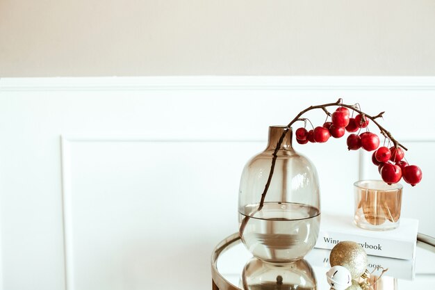 Salón decorado. Mesita de noche con ramo de frutos rojos en florero de vidrio, libro, vela delante de la pared beige