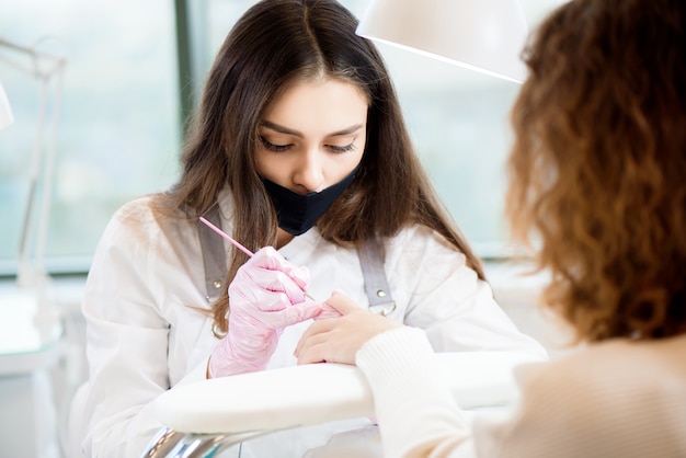 Salón de manicura y pedicura, covid-19 y distancia social. Maestro en guantes de goma y cliente joven en máscara protectora en el interior del estudio de belleza.