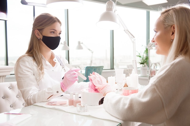 Salón de manicura y pedicura, covid-19 y distancia social. Maestro en guantes de goma y cliente joven en máscara protectora en el interior del estudio de belleza.