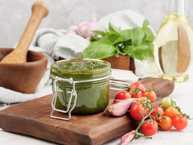 Salsa de pesto de albahaca italiana tradicional en un frasco de vidrio sobre una tabla de madera junto con albahaca y tomates.