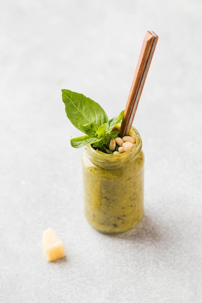 Salsa de pesto casera en un frasco con una hoja de albahaca, piñones, una rodaja de queso parmesano y una cuchara Foto vertical