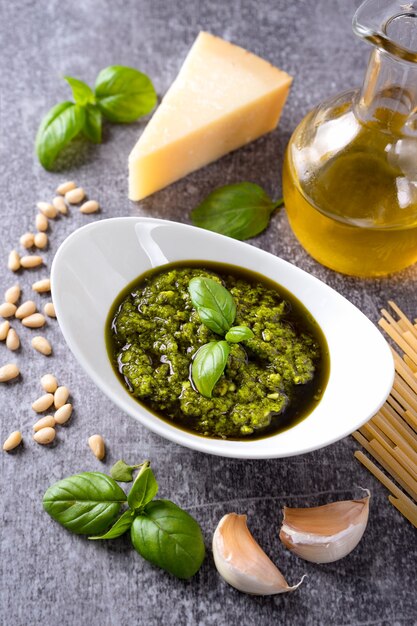 Salsa pesto casera con ingredientes. Salsa de pesto en un tazón blanco con albahaca, aceite de oliva, piñones y queso parmesano sobre fondo de hormigón gris