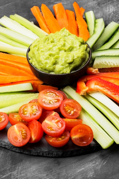 Foto salsa tradicional latinoamericana guacamole en un tazón y diversas verduras (zanahorias, tomates, pepinos, apio) sobre un fondo oscuro.