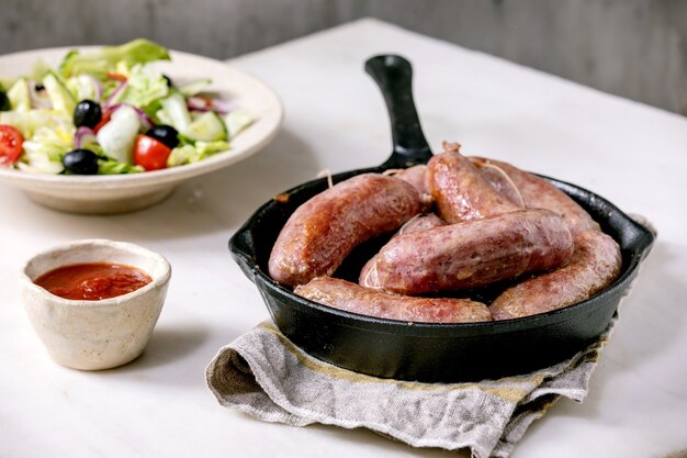 Salsicha de linguiça italiana grelhada em frigideira de ferro fundida, servida com molho de tomate e prato de salada de legumes frescos.