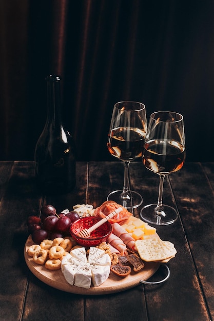 Foto salsicha e queijo cortados em tábua redonda com biscoitos de frutas e molho com dois copos de vinho branco e uma garrafa