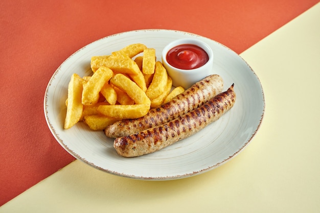 Salsichas clássicas alemãs grelhadas com batatas fritas e molho vermelho em prato branco sobre superfície colorida.