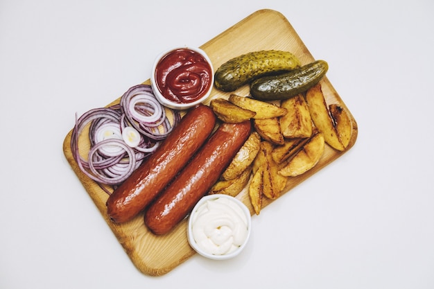 Salsichas com batatas assadas, molhos, cebolas e pepinos em conserva na placa de madeira na vista superior do fundo branco