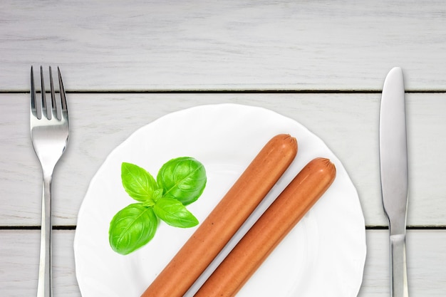 Salsichas de salsicha cozidas saborosas decoradas com folhas de manjericão em um prato com garfo e faca em uma mesa de madeira branca