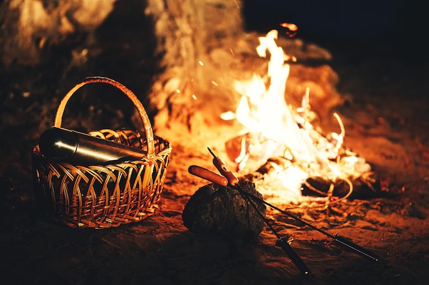 Salsichas em espetos e uma garrafa térmica na cesta contra o fogo à noite.