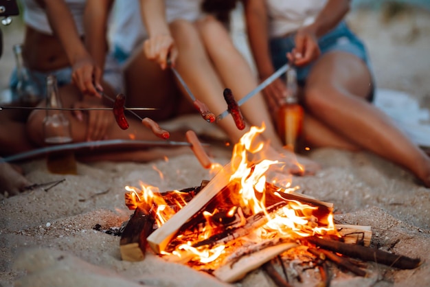 Salsichas fritas na fogueira Grupo de jovens amigos sentados na praia e fritar salsichas Tempo de acampamento