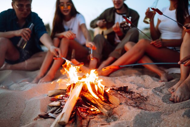 Salsichas fritas na fogueira Grupo de jovens amigos sentados na praia e fritar salsichas Tempo de acampamento