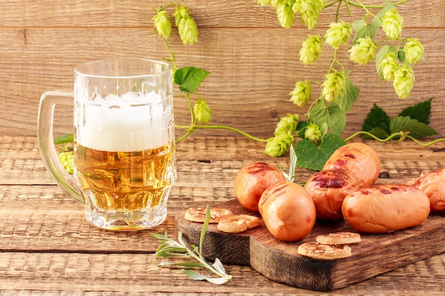 Salsichas fritas na tábua de madeira e copo de cerveja no fundo de madeira com a planta de lúpulo. churrasco, piquenique