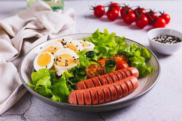 Foto salsichas fritas tomates alface e um sanduíche com um ovo cozido em um prato na mesa