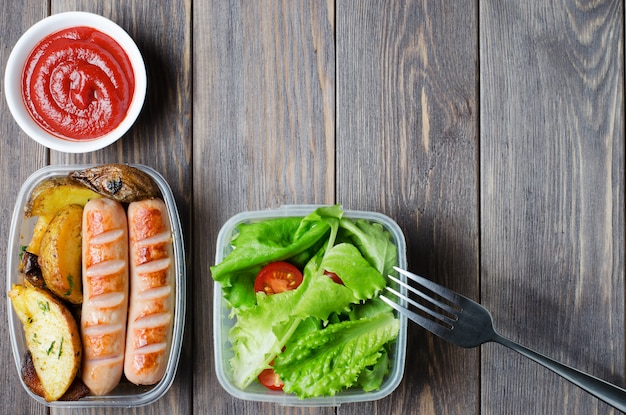 Salsichas grelhadas, batatas, alface verde com tomate em uma caixa de plástico.