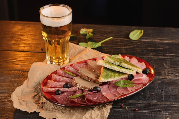 Salsichas sortidas, jamon e presunto com fatias de pão frito num prato vermelho. Em uma mesa de madeira