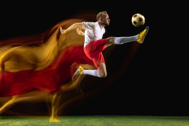 Saltar. Jugador de fútbol o fútbol masculino caucásico joven en ropa deportiva y botas pateando la pelota para el gol en luz mixta en la pared oscura. Concepto de estilo de vida saludable, deporte profesional, afición.