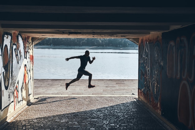 Foto salto em comprimento. comprimento total de um jovem africano em roupas esportivas, correndo enquanto se exercita perto do rio ao ar livre