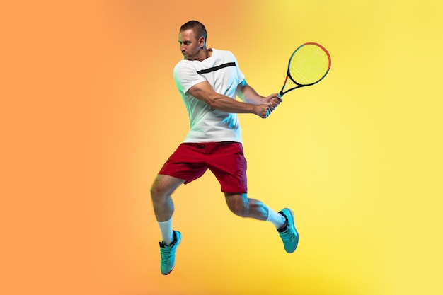 En salto. Un hombre caucásico jugando al tenis en el fondo del estudio con luz de neón. Colocar al joven jugador profesional masculino en movimiento o acción durante el juego deportivo. Concepto de movimiento, deporte, estilo de vida saludable.