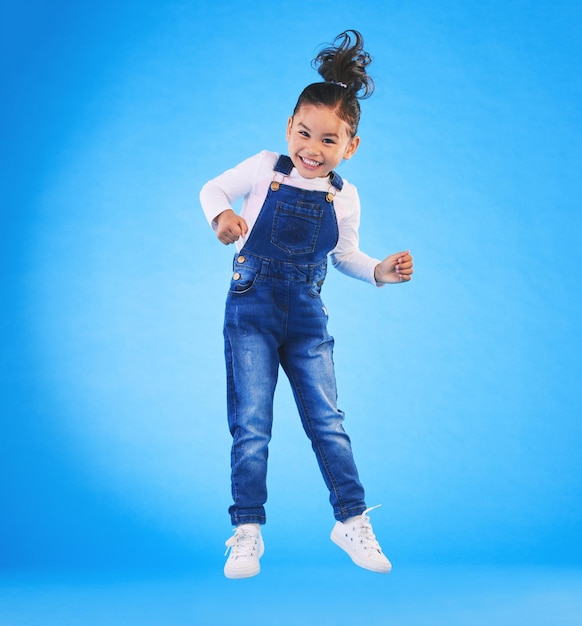 Saltos felices y retrato de un niño en un estudio con emoción, alegría o expresión de sorpresa. Sonrisa de felicidad y longitud completa de una niña modelo moviéndose para recibir buenas noticias aisladas en un fondo azul.