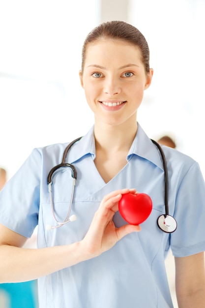 salud, caridad y concepto médico - doctora o enfermera sonriente con corazón y estetoscopio en el hospital con equipo en segundo plano