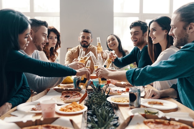 ¡Salud para nosotros! Grupo de jóvenes en ropa casual brindando y sonriendo mientras tienen una cena en el interior