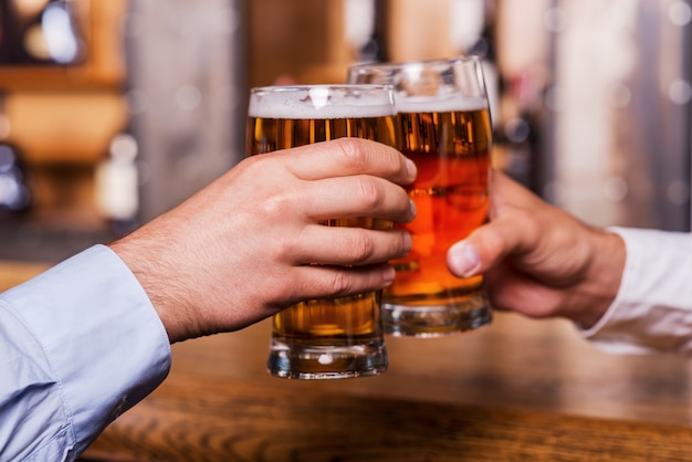 ¡Salud! Primer plano de dos hombres en camisetas brindando con cerveza en la barra del bar