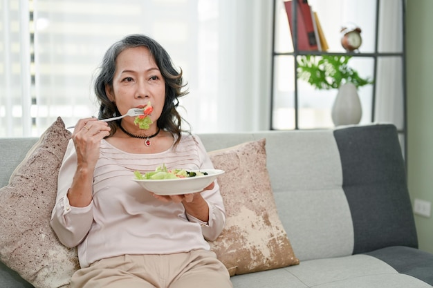 Saludable mujer asiática jubilada de los años 60 comiendo ensalada saludable mientras ve la televisión en su sala de estar