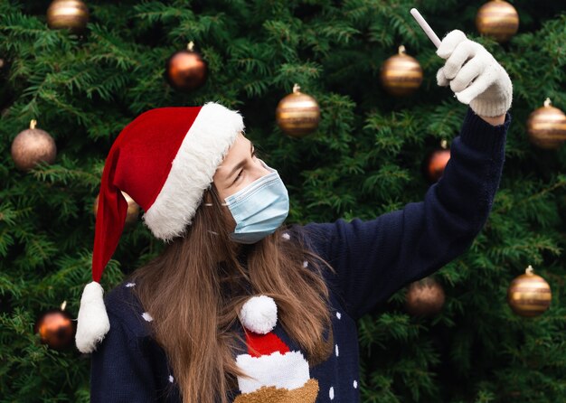 Saludos navideños online. Close up Retrato de mujer con un sombrero de santa claus y una máscara médica con emoción. En el contexto de un árbol de Navidad. Pandemia de coronavirus