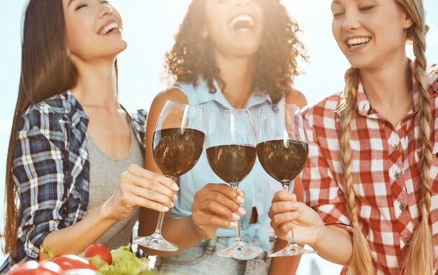 Saludos a tres mujeres jóvenes y felices sosteniendo copas con vino y riendo mientras está de pie en