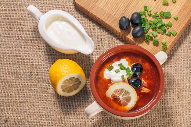 Salzkrautsuppe mit Fleisch, geräucherten Würstchen, Kartoffeln, Tomaten, marinierter eingelegter Gurke, Zitrone, schwarzen Oliven und Sauerrahm in keramischer Suppenschüssel mit Zutaten. Ansicht von oben