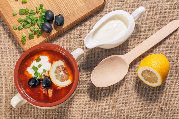 Salzkrautsuppe mit Fleisch, geräucherten Würstchen, Kartoffeln, Tomaten, marinierter eingelegter Gurke, Zitrone, schwarzen Oliven und Sauerrahm in Suppenschüssel aus Keramik, Schneidebrett, Holzlöffel. Ansicht von oben.