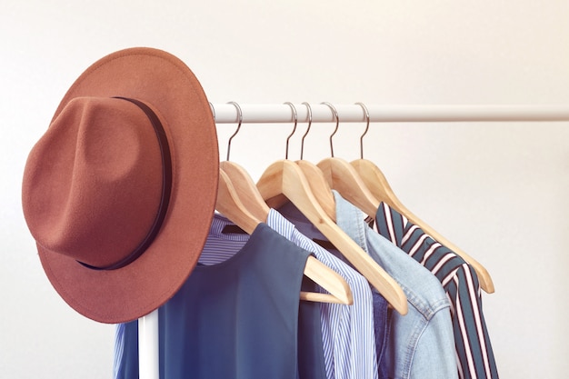 Sammlung Kleidung mit dem braunen Hut, der am Gestell nahe weißer Wand hängt. Kleidung für Frauen in blauen Farben. Büro-Stil.