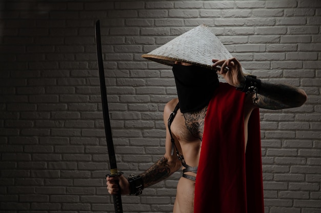 Samurai con una capa roja y un sombrero triangular asiático con tatuajes y una espada katana en las manos contra el fondo de una pared de ladrillos