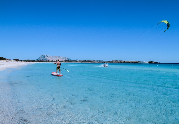 San Teodoro - La Cinta Beach - Juli 2017 - Ein junger Athlet paddelt auf einem Stand-Up-Paddel auf dem kristallklaren Meer Sardiniens, während ein Kitesurfer neben ihm auf dem Wasser rutscht