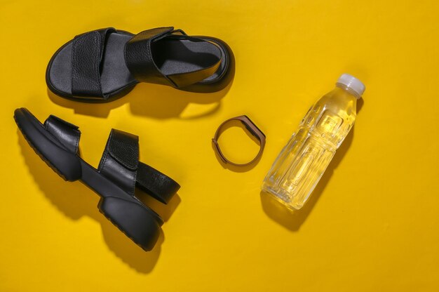 Sandalias, pulsera inteligente y botella de agua sobre un fondo amarillo brillante. Vista superior
