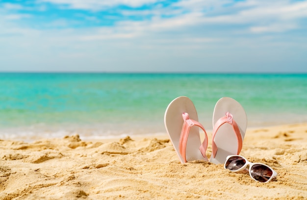 Sandalias rosas y blancas, gafas de sol en la playa de arena en la playa. Chanclas y gafas de estilo casual. Vacaciones de verano en playa tropical. Divertido viaje en la playa de arena. Vibras de verano.