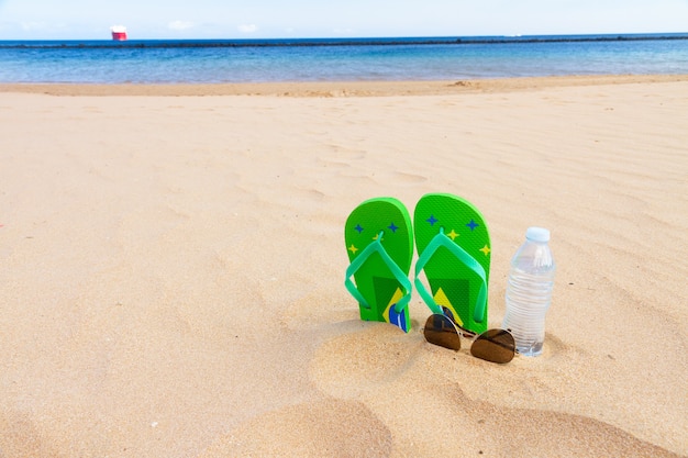 Sandalias verdes en la playa con botella de agua clara y vasos